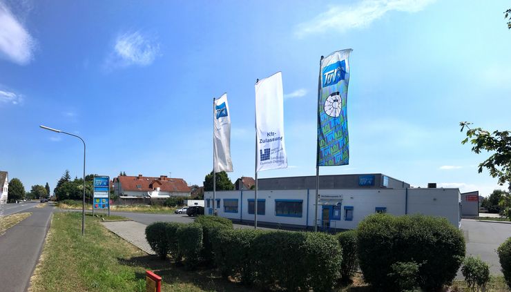 Ein Gewerbegebiet mit drei Fahnen, von denen eine für eine Kfz-Zulassung wirbt, und einem Gebäude mit der Aufschrift "TUEV Hessen". Außerdem gibt es einige Bäume und eine Straße.