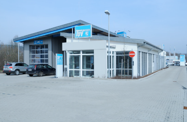 Ein modernes Gebäude mit blau-weißer Fassade und der Aufschrift "TUEV Hessen". Draußen sind Autos geparkt, und die Umgebung scheint ein Gewerbegebiet mit klarem Himmel zu sein.