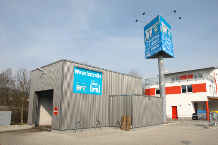Eine modern aussehende Autowaschanlage mit einem blauen Schild mit der Aufschrift 'Waschstrassee' und dem Logo von TUEV Hessen. Daneben befindet sich ein hohes blaues Schild mit verschiedenen Serviceangeboten für Autos.
