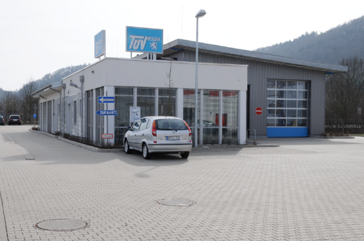 Ein modernes Autohaus mit einem weißen Auto, das vor dem Ausstellungsraum geparkt ist. Das Autohaus hat ein blaues Schild mit der Aufschrift "TUEV Hessen" und ein weiteres Schild mit der Aufschrift "ZUFAHRT". Die Umgebung besteht aus einem Parkplatz und einer bergigen Kulisse.
