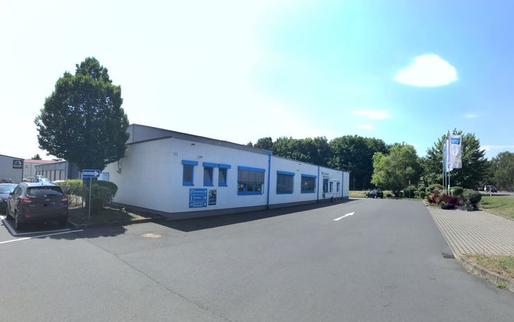 Ein weißes Gebäude mit blauen Akzenten, das sich auf einem Parkplatz befindet, auf dem ein paar Autos parken. Im Hintergrund sind Bäume und ein klarer blauer Himmel zu sehen.